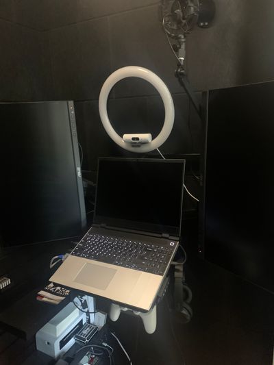 A wall-mounted laptop flanked by a pair of vertical-oriented displays. Above it is a ring light with central webcam and microphone boom folded up and away. Headphones hang on a hook mounted to the right side of the laptop's tray, and a game controller is in a holder mounted to the front right corner of the tray.