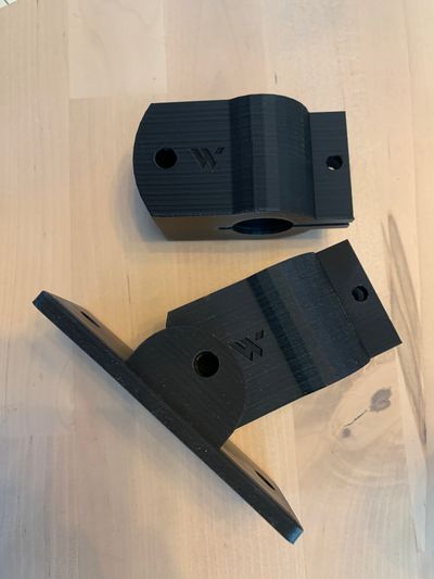 A pair of black, hinged, 3D printed wall mounts sitting on a table unassembled. There is a W embossed on the sides.